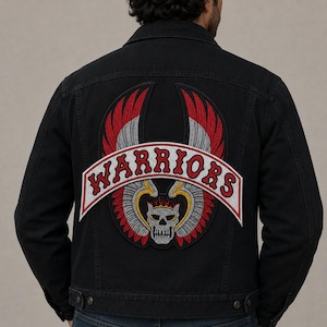 May include: A black denim jacket featuring a large embroidered patch on the back. The patch displays a central skull with flames, surrounded by stylized wings in red, white, and grey. A curved banner above the skull reads WARRIORS in red.