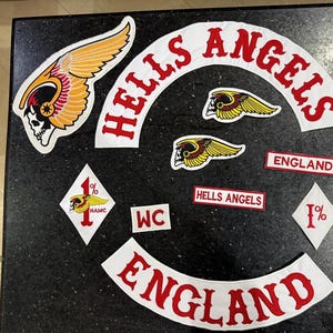 Könnte beinhalten: Eine Sammlung bestickter Aufnäher mit dem Hells Angels Logo, darunter ein großer geflügelter Totenkopf, geschwungene Schriftzüge mit HELLS ANGELS und ENGLAND, zwei rautenförmige 1%-Aufnäher sowie kleinere rechteckige Aufnäher mit Text.