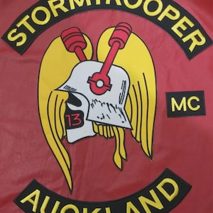 May include: A red patch featuring a central graphic of a white skull wearing a helmet with yellow wings and two red pistons. The text STORMTROOPER is arched above, AUCKLAND is arched below, and MC is on the right, all in yellow letters on black.