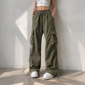 May include: Olive green cargo pants featuring a high-waisted elastic waistband with a drawstring, two large side cargo pockets with flaps, and long decorative fabric ties hanging from the pockets. The pants have a loose, wide-leg fit.