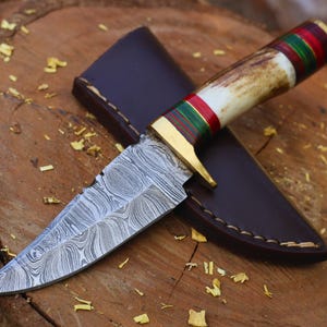Puede incluir: Un cuchillo de hoja fija con una hoja de acero Damasco estampada, una guarda de latón y un mango de asta con espaciadores rojos y verdes. El cuchillo viene acompañado de una funda de cuero marrón oscuro con costuras visibles en el borde.