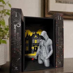 Pode incluir: Um inserto de estante com uma figura de torso masculino cinza impressa em 3D com moletom e uma pequena rosa vermelha, posicionada entre dois livros Haunting Adeline de H.D. Carlton com uma ilustração de uma casa ao fundo.