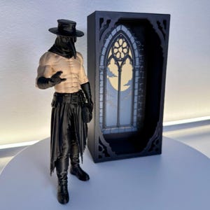 Op de afbeelding: Een pestmeester-actiefiguur met een gespierd bovenlichaam, een zwarte hoed met brede rand, een vogelachtig masker, handschoenen en een rafelige rok. Daarnaast staat een zwart rechthoekig frame met een gotisch boogvenster en een maanverlichte lucht.
