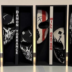 May include: Four black rectangular display boxes, each containing two stylized masks with distinct graphic patterns. The boxes feature vertical dividers labeled with the names Michael Crist, Damon Torrance, Will Grayson, and Kai Mori in bold text.