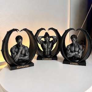 Op de afbeelding: Drie donkergrijze, gespierde mannelijke bustes omlijst door gebogen, vleugelachtige structuren. Elk figuur staat op een rechthoekige voet met gouden letters: Rhysand, Cassian en Azriel. De figuren hebben gedetailleerde anatomische kenmerken.