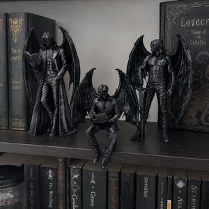 Op de afbeelding: Drie donkergrijze, getextureerde gevleugelde humanoïde figuren. De linkerfiguur staat een boek te lezen, de middelste figuur zit lezend op de rand van een plank en de rechterfiguur staat met een zwaard. Alle drie hebben grote, gedetailleerde vleugels.