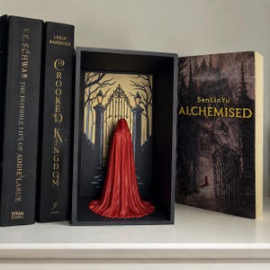 Pode incluir: Um inserto de estante apresentando uma figura 3D de capuz vermelho em frente a uma ilustração de um portão de ferro preto. O inserto está alojado em uma moldura retangular preta, posicionada entre dois livros pretos e o livro Alchemised de SenLinYu.