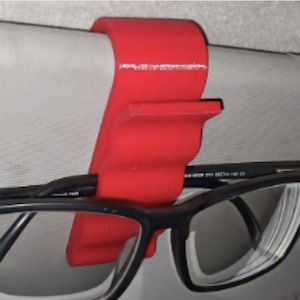 Könnte beinhalten: Ein leuchtend roter, U-förmiger Kunststoffclip zur Befestigung an einer Sonnenblende im Fahrzeug. Der Clip verfügt über eine kleine horizontale Ablage, die eine Brille mit schwarzem Rahmen sicher an ihrem Platz hält und griffbereit macht.