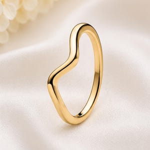 May include: A polished gold ring featuring a smooth, continuous band with a distinct wave-like curve along one side. The metal surface is reflective and uniform in color, showcasing a simple, rounded profile that maintains a consistent thickness.