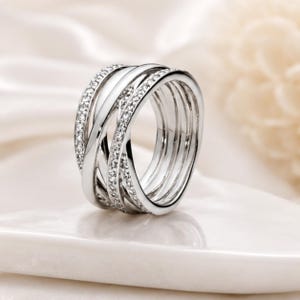 May include: A silver-toned ring featuring a multi-band crossover design. The band consists of several intertwined strands, with two of the outer strands encrusted with small, clear, sparkling stones that catch the light along the entire surface.