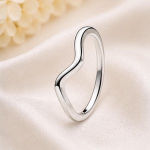 Puede incluir: Un anillo de plata pulida que presenta un contorno distintivo en forma de V. La banda tiene una superficie metálica lisa y reflectante con un diseño fluido y continuo que crea una sutil punta a lo largo del borde superior de la pieza.
