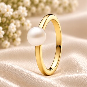 Op de afbeelding: Een gouden ring met een enkele, gladde, ronde witte parel aan de bovenkant van de band. De band heeft een gepolijste, reflecterende gouden afwerking en de parel is gepositioneerd als een opvallend middelpunt op de ronde metalen structuur.