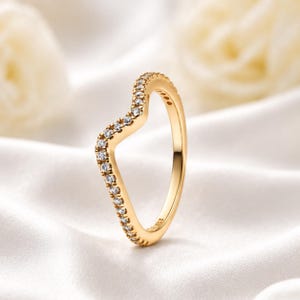 Puede incluir: Un anillo de tono dorado con un contorno distintivo en forma de V. La superficie exterior de la banda está engastada con una fila continua de pequeñas piedras claras de corte redondo que siguen la silueta angular de la pieza de joyería.