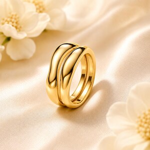 May include: A polished gold ring featuring a double-band design with smooth, rounded surfaces. The ring has a high-shine metallic finish that reflects light, creating bright highlights along the curved edges of the two connected bands.