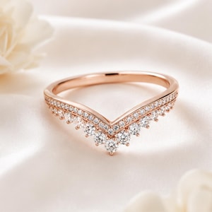 May include: A rose gold chevron ring featuring a V-shaped band adorned with a row of small pavé-set clear stones and a series of larger round-cut clear stones along the bottom edge, creating a layered, sparkling appearance on the metal band.