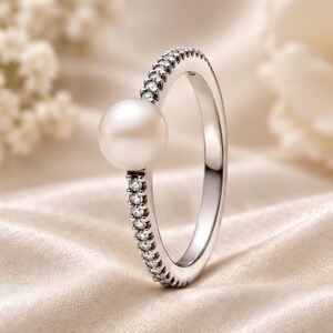 Op de afbeelding: Een zilveren ring met een enkele ronde witte parel aan de bovenkant, waarbij de band is versierd met een rij kleine, heldere, fonkelende steentjes langs de buitenrand, wat dit sieraad een gepolijste en reflecterende uitstraling geeft.