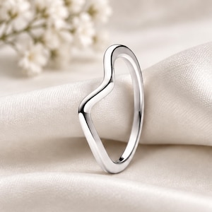 May include: A polished silver-toned ring featuring a distinct V-shaped contour. The band has a smooth, reflective metallic surface with a continuous, fluid design that creates an asymmetrical silhouette, showcasing a clean and simple aesthetic.