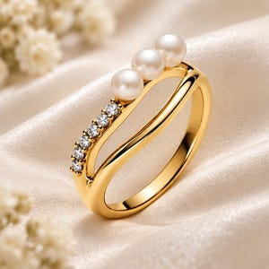 Puede incluir: Un anillo de tono dorado con un diseño de banda dividida, que presenta una hilera de pequeños cristales transparentes en un lado y tres perlas blancas esféricas dispuestas en línea a lo largo de la curva superior de la banda.