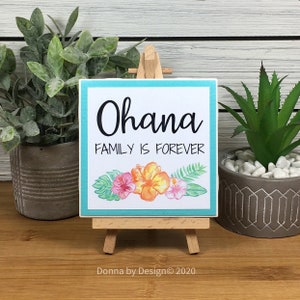 May include: A small wooden sign with a turquoise border and a white background. The sign says "Ohana Family is Forever" in black text. The sign is decorated with a watercolor illustration of tropical flowers and leaves. The sign is displayed on a small wooden easel.