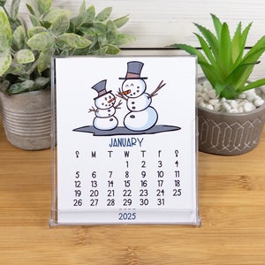 May include: A white desk calendar with a January 2025 calendar grid. The calendar features a cartoon illustration of two snowmen with black top hats and carrot noses. The snowmen are standing on a blue background.