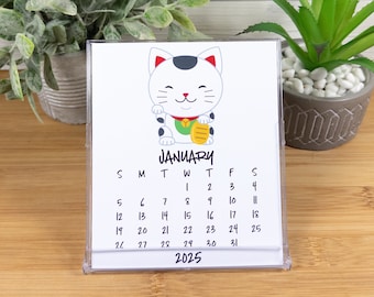 May include: A white desk calendar with a cartoon image of a lucky cat holding a gold coin. The calendar is for January 2025 and has the days of the week labeled.