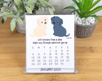 May include: A white desk calendar with a January 2025 calendar grid. The calendar features a cartoon illustration of two Labrador puppies, one yellow and one black. The text "Life lessons from a dog: Make new friends wherever you go" is printed below the puppies.