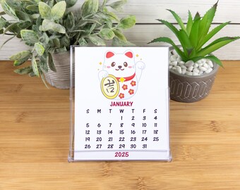 May include: A white desk calendar with a cartoon illustration of a Japanese Maneki-Neko, or lucky cat, holding a gold coin with the Japanese character for "lucky" on it. The calendar is for January 2025 and features a red and white floral design.