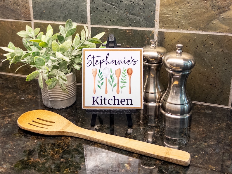 K&ouml;nnte beinhalten: Ein kleines Holzschild mit dem Text "Stephanie's Kitchen" in lila Schrift. Das Schild ist mit einer Aquarell-Illustration von K&uuml;chenutensilien und Gr&uuml;npflanzen verziert. Das Schild steht auf einem kleinen Staffelei auf einer schwarzen Arbeitsplatte.