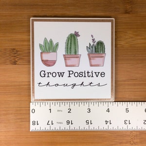 May include: White square coaster with a brown border. The coaster features a hand-drawn illustration of three potted cacti with the text "Grow Positive thoughts" in black.