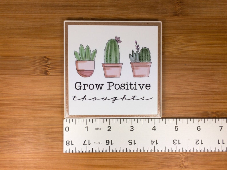 May include: White square coaster with a brown border. The coaster features a hand-drawn illustration of three potted cacti with the text "Grow Positive thoughts" in black.