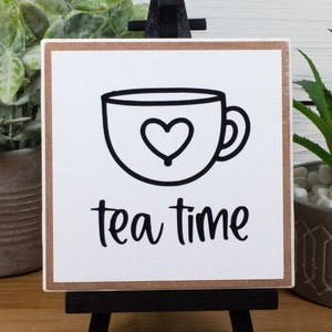 May include: A white wooden sign with a black outline of a teacup with a heart inside and the text "tea time" in black cursive lettering.