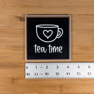 May include: Black square sign with white text and a white outline of a teacup with a heart in it. The text reads "tea time".