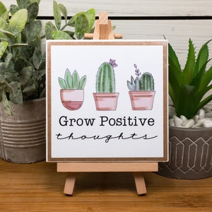 May include: A wooden sign with a white background and a brown frame. The sign has a hand-drawn illustration of three potted plants, two cacti and one succulent. The text on the sign reads "Grow Positive thoughts".