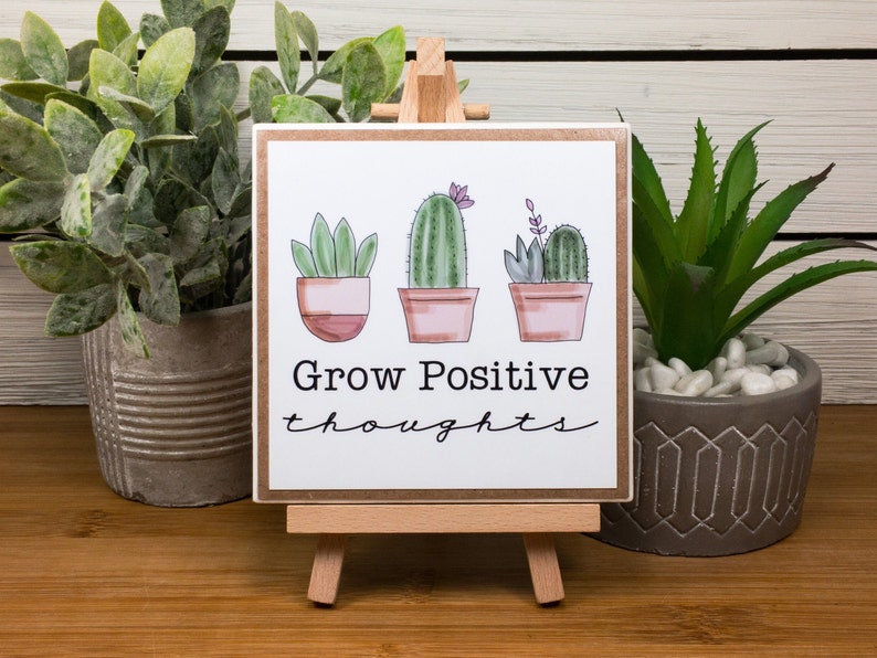 May include: A wooden sign with a white background and a brown frame. The sign has a hand-drawn illustration of three potted plants, two cacti and one succulent. The text on the sign reads "Grow Positive thoughts".