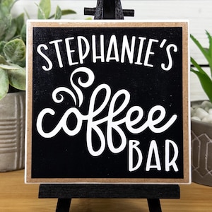 May include: A black wooden sign with a white border and the text "Stephanie's Coffee Bar" in white cursive font.