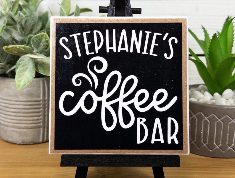 May include: A black wooden sign with a white border and the text "Stephanie's Coffee Bar" in white cursive font.