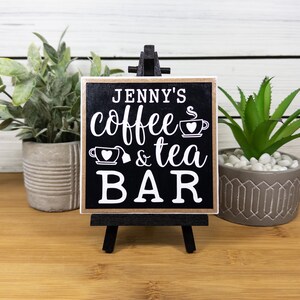 May include: A black wooden sign with a gold border reads "Jenny's coffee & tea bar" with a coffee cup and teacup graphic. The sign is on a small easel.