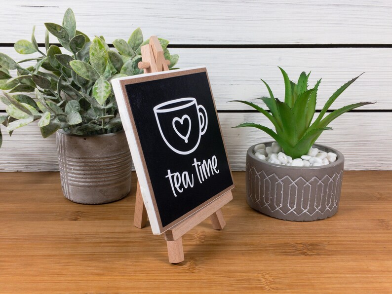 May include: A small black chalkboard sign with white text and a white outline of a teacup with a heart inside. The sign reads "tea time" and is displayed on a small wooden easel. The sign is sitting on a wooden table with a green plant in a gray pot on either side.