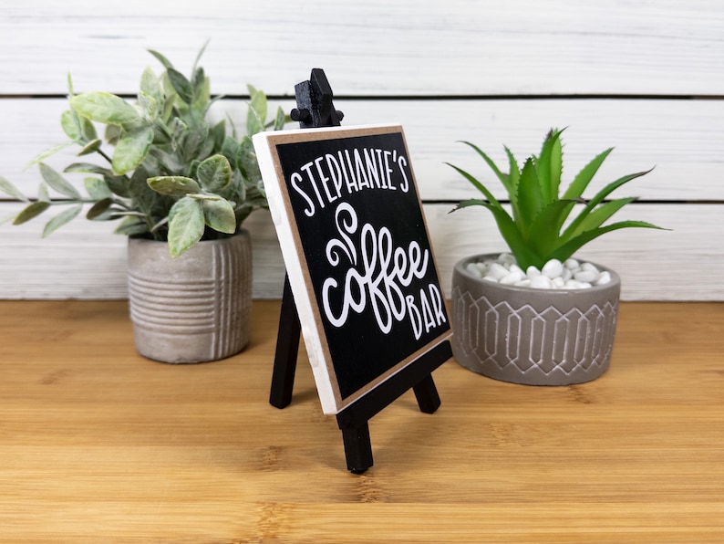 May include: A black chalkboard sign with a white easel stand. The sign says "Stephanie's Coffee Bar" in white lettering.