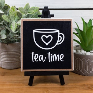 May include: Black wooden sign with a white outline of a teacup with a heart inside and the text "tea time". The sign is on a small easel.