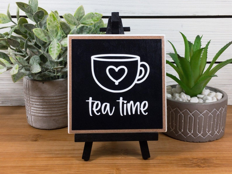 May include: Black wooden sign with a white outline of a teacup with a heart inside and the text "tea time". The sign is on a small easel.