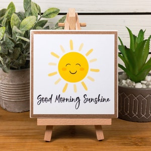 May include: A small wooden easel displaying a white square sign with a yellow smiling sun and the text "Good Morning Sunshine". The sign is on a wooden table with two potted plants.