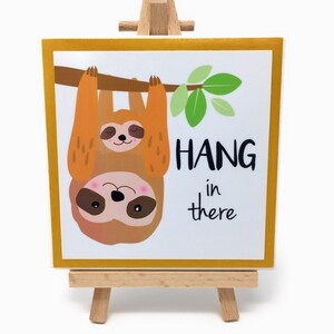Puede incluir: Una impresi&oacute;n art&iacute;stica cuadrada sobre un peque&ntilde;o caballete de madera. La impresi&oacute;n presenta un perezoso de dibujos animados colgando de una rama con las palabras "HANG in there" en texto negro. El perezoso es marr&oacute;n y naranja con una cara rosa.