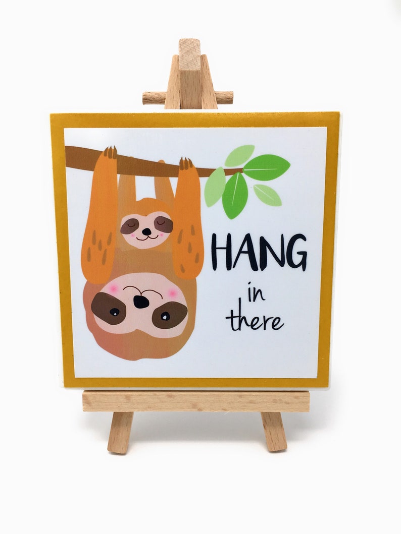 Puede incluir: Una impresi&oacute;n art&iacute;stica cuadrada sobre un peque&ntilde;o caballete de madera. La impresi&oacute;n presenta un perezoso de dibujos animados colgando de una rama con las palabras "HANG in there" en texto negro. El perezoso es marr&oacute;n y naranja con una cara rosa.
