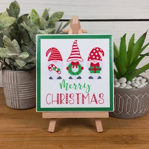 Könnte beinhalten: Eine weiße Keramikfliese mit grünem Rand zeigt drei Gnome, die rote und weiße Hüte tragen. Die Gnome halten einen Zuckerstock, einen Kranz und eine Geschenkbox. Die Fliese trägt die Aufschrift "Merry Christmas" in roten und weißen Buchstaben.