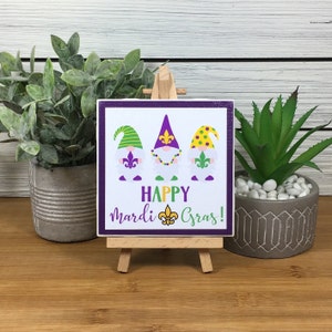 Könnte beinhalten: Ein quadratisches Schild mit drei Zwergenillustrationen, die lila, grüne und gelbe Hüte tragen und jeweils eine Lilie halten. Auf dem Schild steht "HAPPY Mardi Gras!" in lila und grünen Buchstaben, das auf einer kleinen Holzstaffelei ausgestellt ist.