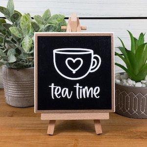 May include: Black chalkboard sign with a white teacup and heart design. The sign reads "tea time" and is displayed on a small wooden easel.