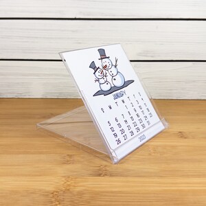 May include: A clear plastic desk calendar with a snowman illustration. The calendar is for January 2025 and shows the days of the week and dates.