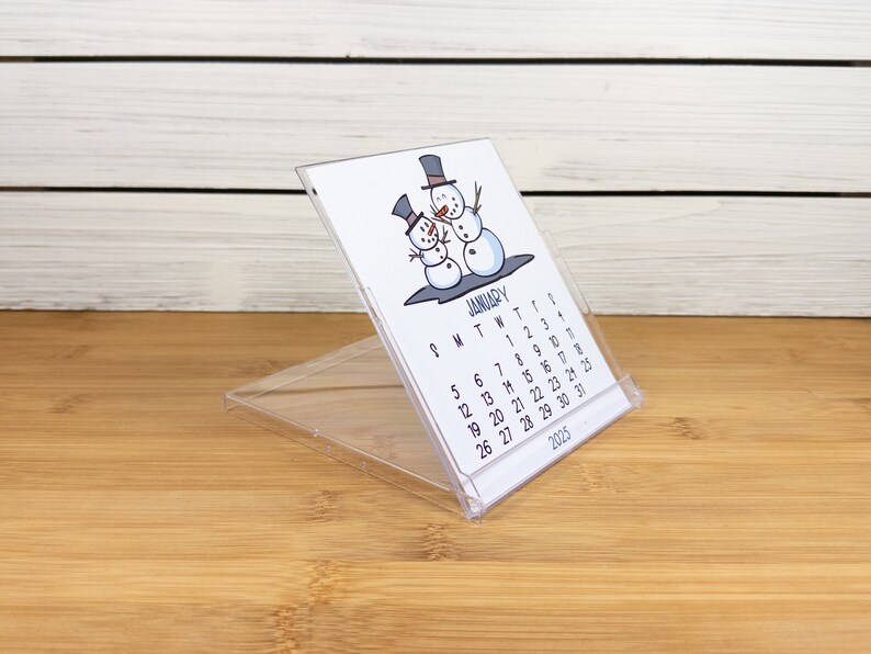 May include: A clear plastic desk calendar with a snowman illustration. The calendar is for January 2025 and shows the days of the week and dates.