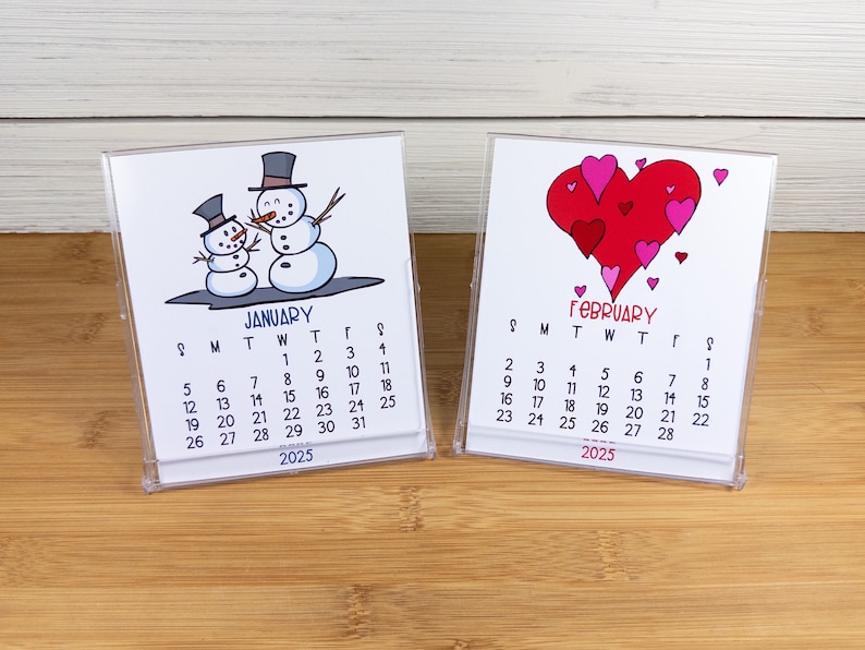May include: Two small desk calendars with a winter and a Valentine's Day theme. The January calendar features a cartoon illustration of two snowmen. The February calendar features a cartoon illustration of a large red heart with smaller hearts around it. Both calendars are for the year 2025.